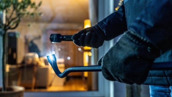 Ein Einbrecher späht von der Gartenseite her eine Wohnung aus, hält Werkzeug und Taschenlampe in der Hand. © picture alliance Foto: Jochen Tack