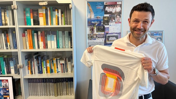 Der Arzt Caspar Grim steht in seinem Büro und hält ein T-Shirt der deutschen Olympiamannschaft in seinen Händen. © NDR Foto: Marlena Maerz
