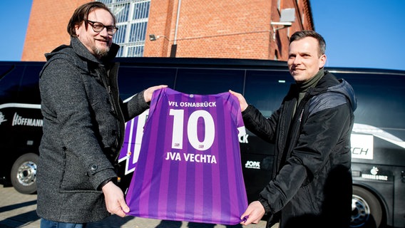 Michael Welling, Geschäftsführer des VfL Osnabrück, und Sebastian Rüther, Pressesprecher des VfL Osnabrück, stehen mit einem Trikot des Vereins im Innenhof der Justizvollzugsanstalt für Jungtäter. © dpa-Bildfunk Foto: Hauke-Christian Dittrich/dpa