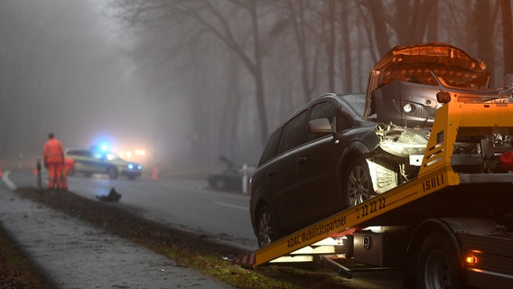 Im Vordergrund zieht ein Abschleppauto einen beschädigten Wagen auf die Transportfläche. Im Hintergrund steht ein Polizeiwagen mit eingeschaltetem Blaulicht quer auf der Straße. © dpa-Bildfunk Foto: Lars Penning/dpa