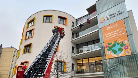 Als Superhelden verkleidete Feuerwehrleute erklimmen über eine Drehleiter den Balkon im Kinderkrankenhaus in Osnabrück, um Geschenke zu überreichen. © NDR Foto: Andreas Gervelmeyer