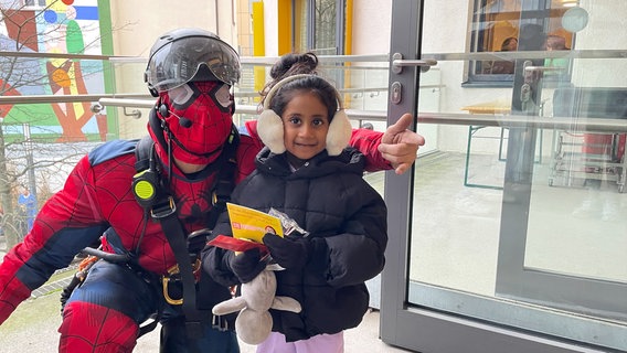 Ein als Spiderman verkleideter Feuerwehrmann überreicht einem kleinen Mädchen auf dem Balkon des Kinderkrankenhauses in Osnabrück ein Geschenk. © NDR Foto: Andreas Gervelmeyer