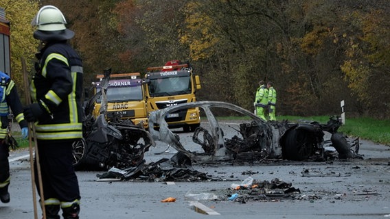 Auf einer Landstraße stehen zwei Abschleppwagen und mehrere Rettungskräfte in neongelber Kleidung. Im Vordergrund sind völlig ausgebrannte Reste eines Autos zu sehen. © picture alliance Foto: Michel Fritzemeier