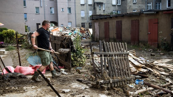 Ein Mann in Gummistiefeln schaufelt Matsch in einem Innenhof, in dem viel zerbrochenes Holz und Müll liegen. - In Polen haben Überschwemmungen große Zerstörung angerichtet. © picture alliance / ZUMAPRESS.com | Attila Husejnow Foto: Attila Husejnow