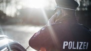 Ein Polizist in Uniform steht bei Gegenlicht neben einem Polizeiauto und hält einen Telefonhörer in der Hand. © NDR Foto: Julius Matuschik