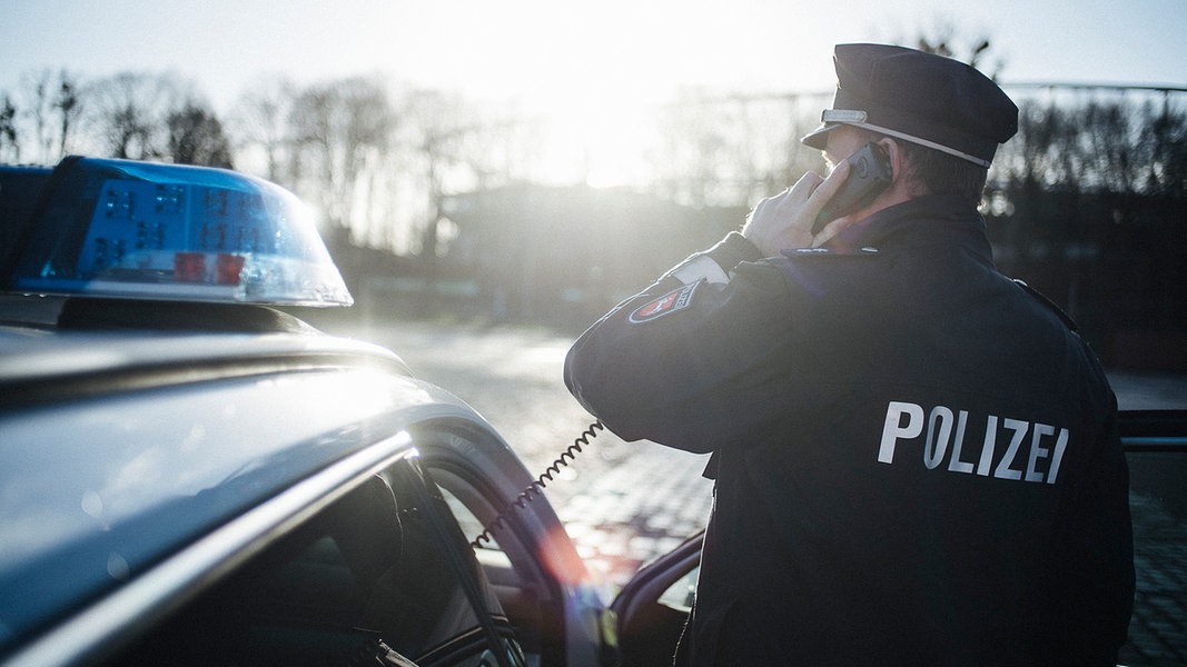 Ein Polizist in Uniform steht neben einem Polizeiauto und hält einen Telefonhörer in der Hand.