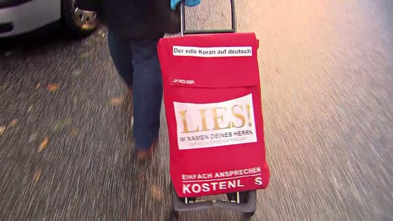 Jemand zieht einen Rollkoffer mit der Aufschrift "Lies!". © NDR 