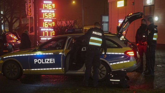 Drei Polizisten stehen vor einem Polizeiwagen. Im Hintergrund zu sehen ist die Preistafel einer Tankstelle. © picture alliance/ABBfoto Foto: ABBfoto