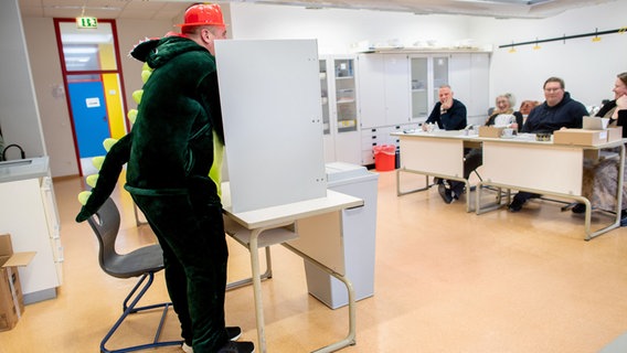 Ein als Dinosaurier kostümierter Wähler steht in einem Wahllokal bei der Stimmabgabe hinter einem Sichtschutz. © dpa Foto: Hauke-Christian Dittrich