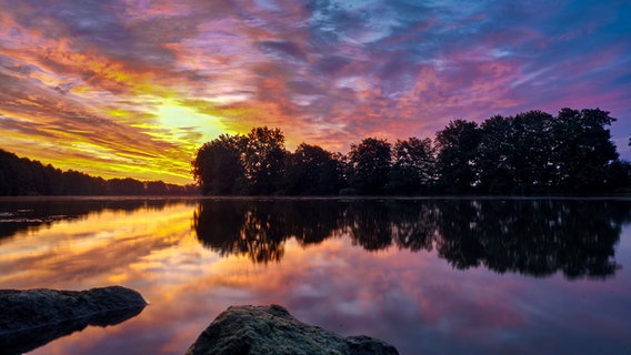 Wolken uns Bäume spiegeln sich in einem Gewässer, am Himmel zeigen sich gelbe, rot und lila Farben. © NDR Foto: Christian Ravenstein