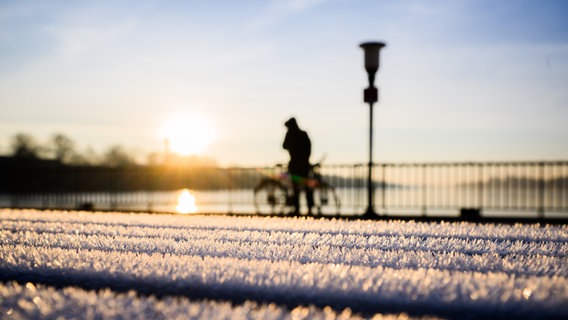 Ein Mann hat sein Fahrrad bei Sonnenaufgang vor dem Maschsee abgestellt. Auf einer Bank liegt Raureif. © picture alliance/dpa Foto: Julian Stratenschulte