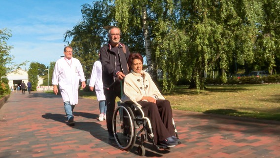 Ein Ehemann rollt seine kranke Frau im Rollstuhl, hinter ihnen gehen zwei Ärzte in weißen Kitteln. © NDR 