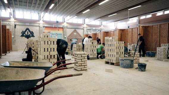 In einer Halle bauen Auszubildende an Mauerwerk. © NDR Foto: Lisa Pandelaki