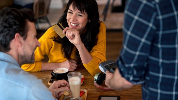 Eine lachende Frau sitzt am Tisch im Café mit Kreditkarte in der Hand, im Vordergrund steht ein Mann mit Kartenlesegerät. © picture alliance / Westend61 | Joseffson Foto: Joseffson