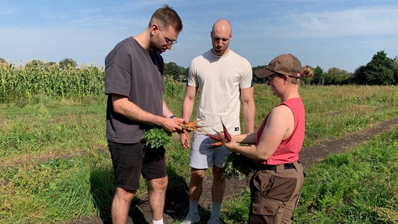 Maik und Marco Möller stehen einer Frau gegenüber und halten frisch geerntete Möhren in den Händen. © NDR Foto: Lena Haamann