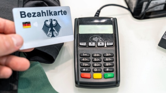 Jemand hält eine guthabenbasierte Debitkarte mit der Aufschrift "Bezahlkarte" vor ein Kartenlesegerät. © IMAGO / Bihlmayerfotografie 