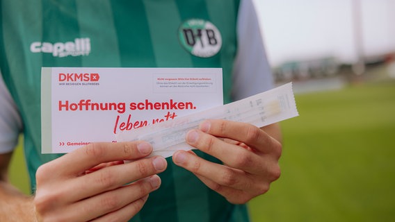 Eine Person in einem Trikot des VfB Lübeck hält ein Registrierungsset der DKMS in die Kamera © Agentur 54 °/DKMS Foto: Agentur 54 °/DKMS