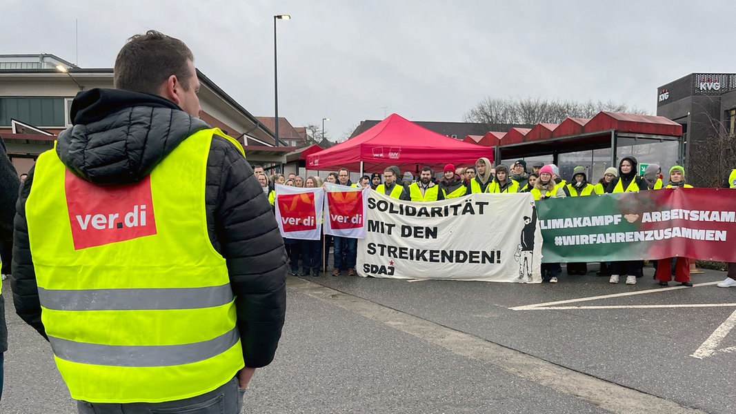 Ein Mann trägt eine gelbe Ver.di-Weste. Im Hintergrund mehrere Streikende mit Plakaten.