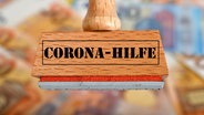 Ein Stempel mit der Aufschrift "Corona-Hilfe", im Hintergrund liegen viele Geldscheine. © picture alliance / Eibner-Pressefoto Foto: Weber/ Eibner-Pressefoto