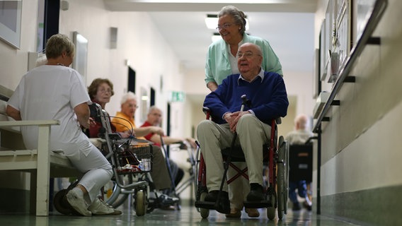 Eine Frau von den "Grünen Damen" schiebt einen alten Mann im Rollstuhl durch einen Krankenhausflur. © picture alliance / dpa Foto: Christian Charisius