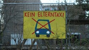 Am Zaun einer Schule hängt ein Schild mit einem durchgestrichenen Auto und der Aufschrift "Elterntaxi". © IMAGO / Hanno Bode Foto: Hanno Bode