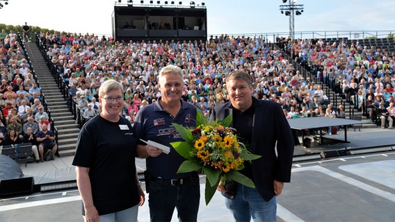 Zwei Männer und eine Frau stehen vor einer menschengefüllten Tribüne bei den Eutiner Festspielen. © ef/Achim Krauskopf 