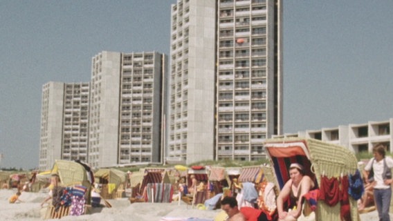 Im Vordergrund sitzen Menschen am Strand, im Hintergrund stehen drei Hochhäuser © NDR Foto: NDR Screenshots