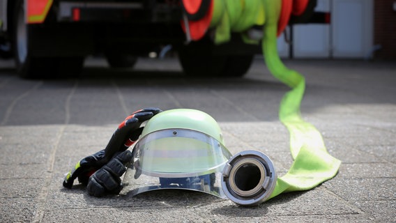 Ein Schutzhelm und Einsatzhandschuhe liegen auf dem Boden neben dem ausgerollten Schlauch eines Feuerwehrautos. © NDR Foto: Pavel Stoyan