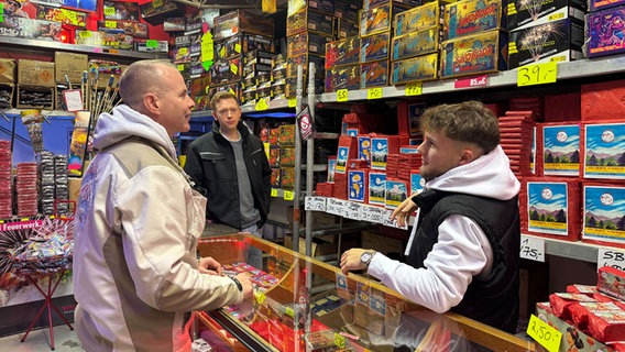 Drei Männer stehen vor gefüllten Regalen in einem Geschäft, in dem Feuerwerk verkauft wird. © NDR Foto: Daniela Tabarelli