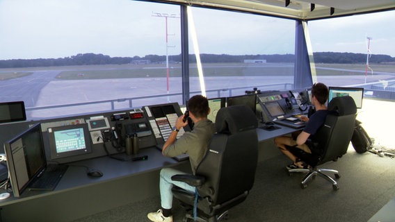 Von hinten sieht man zwei junge Männer in der Kontrollzentrale des Lübecker Flughafen sitzen, die Landebahnen sind ihre Aussicht. © NDR Foto: Lilli Michaelsen