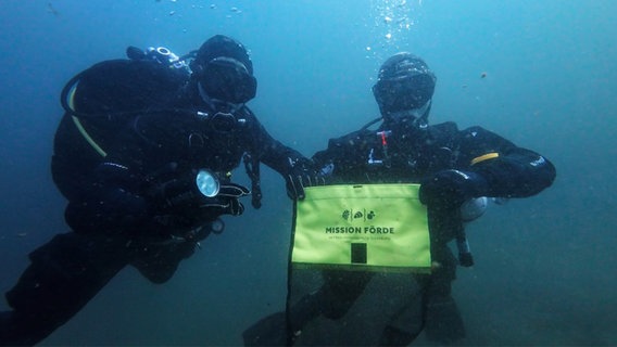 Zwei Taucher unter Wasser, der rechte hält einen Beutel in der Hand, auf dem "Mission Förde" steht. © NDR Foto: Lauritz Graf Bülow von Dennewitz