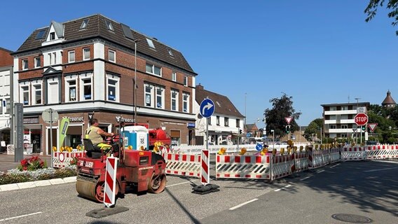 Aufgrund von Straßenarbeiten am Wulf-Isebrand-Platz in Heide sind Umleitungsschilder eingerichtet sowie Absperrungen eingerichtet. Zudem sitzt ein Mann auf einer Walze, um Aspahltierungsarbeiten vorzunehmen. © NDR Foto: NDR