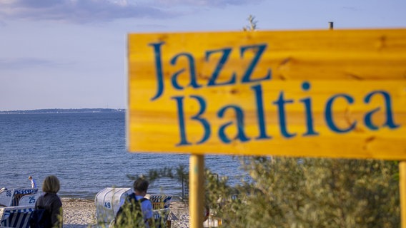 Im Vordergrund ein Schild, auf dem in blauen Lettern "Jazz Baltica" steht, im Hintergrund Strandkörbe und Meer. © JazzBaltica / John Garve_Agentur 54° 