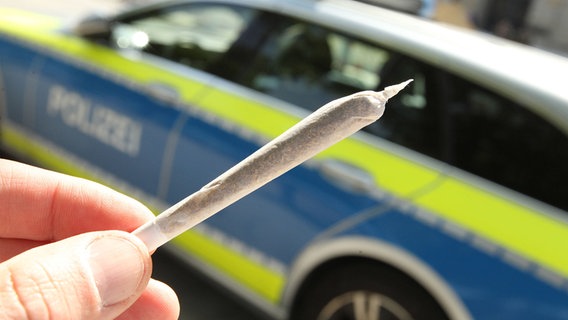 Ein Mann hält einen Joint in der Hand. Im Hintergrund ist ein Einsatzwagen. © IMAGO Foto: Hanno Bode