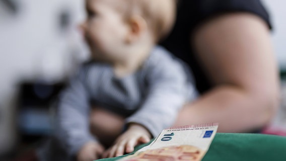 Ein 10-Euro-Schein liegt auf einem Tisch. Im Hintergrund sitzt ein Baby auf dem Schoß einer Frau. © IMAGO / photothek 