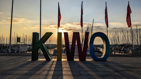 Ein Sonnenaufgang in Kiel-Schilksee. Im Gegenlicht sind die Buchstaben KIWO als Werbemittel am Kai platziert. © Landeshauptstadt Kiel Foto: Gotlind Gädeke