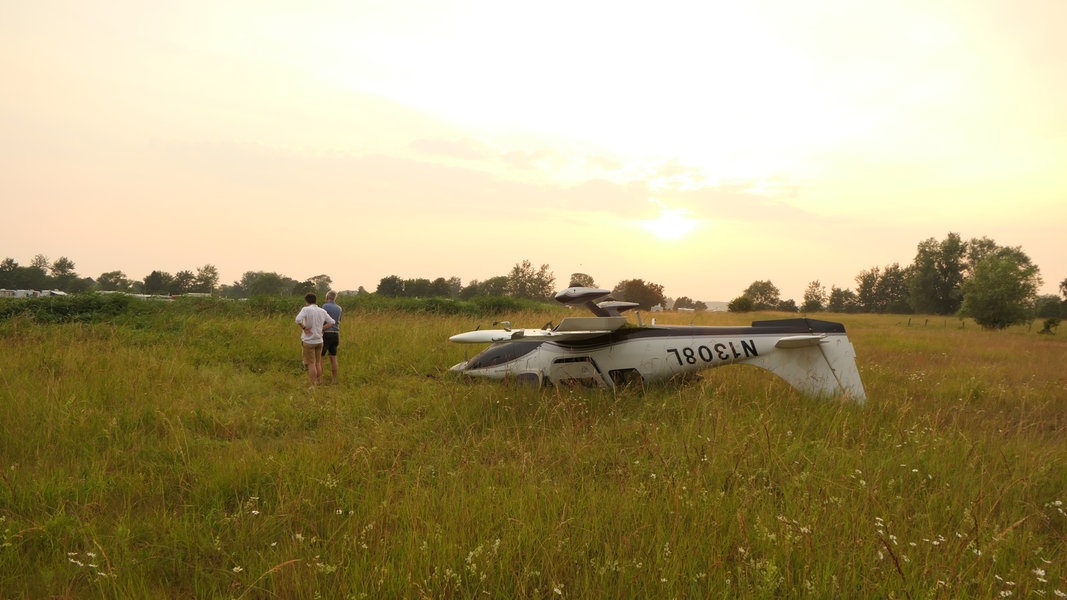 Ein Leichtflugzeug liegt auf dem Rücken auf einer Wiese, zwei Personen stehen daneben. 