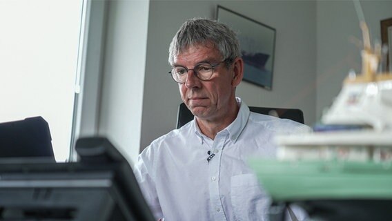 Ein Mann mit Brille sitzt vor seinem Schreibtisch, im Vordergrund ist ein Telefon und ein kleines, türkis-weißes Schiff. © NDR Foto: Dominik Dührsen