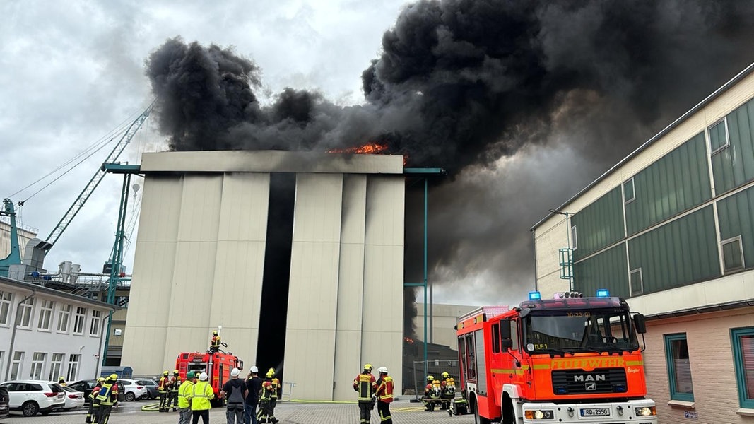 Einsatzkräfte der Feuerwehr stehen vor der brennenden Halle der Lürssen-Werft in Schacht-Audorf.
