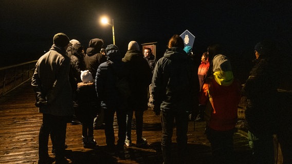 Eine Gruppe steht im Dunkeln auf einem Holzsteg um einen jungen Mann herum. © NDR Foto: Lisa Pandelaki