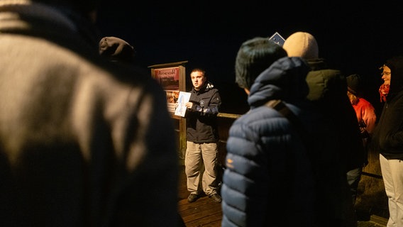 Eine Gruppe steht im Dunkeln auf einem Holzsteg um einen jungen Mann herum. © NDR Foto: Lisa Pandelaki