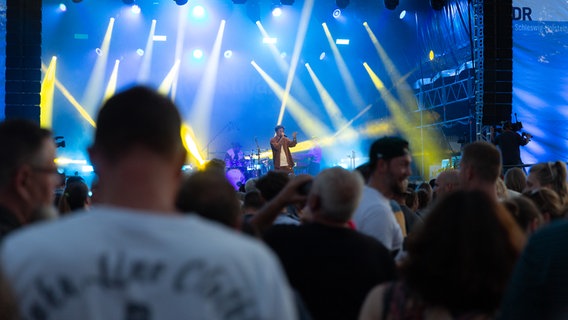 Mehrere Menschen von hinten die vor einer Bühne mit blauem Hintergrund stehen auf der ein Sänger in einem braunen Hemd singt. © NDR Foto: Dominik Dührsen