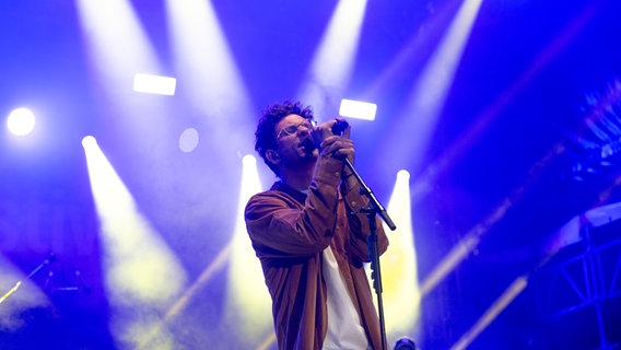 Sänger in einem braunen Hemd steht auf einer Bühne vor blauem Hintergrund. © NDR Foto: Dominik Dührsen