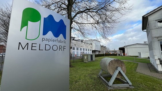 Eine Tafel mit dem Logo der Papierfrabrik Meldorf sowie eine Papierrolle auf einem speziellen Stönder stehen auf dem Rasen auf dem Gelände der Papierfabrik in Tornesch. © NDR Foto: Christoph Deuschle