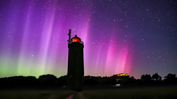 Ein Leuchtturm ragt zwischen Bäumen in den Himmel und ist zwischen den bunten Polarlichtern zu sehen. © Michaela Peter Foto: Michaela Peter