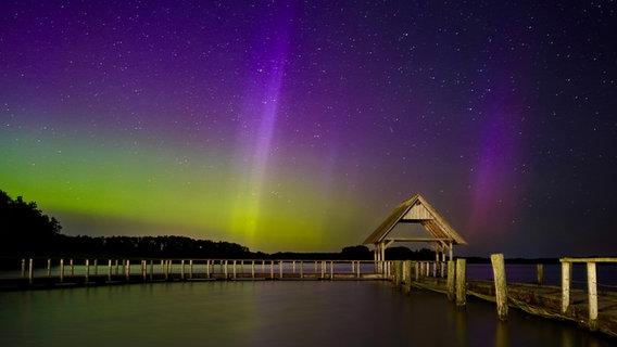 Ein Steg mit einer Hütte wird von goldfarbenen Polarlichtern angeschienen. © Laurens Jürgensen Foto: Laurens Jürgensen