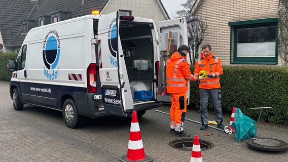 Zwei Mitarbeiter von der Stadtentwässerung Wedel stehen an einem Kanalschacht mit einer Köderbox in den Händen. © NDR Foto: Lena Haamann