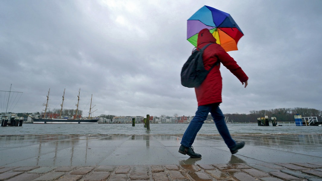 Eine Person geht mit einem bunten Regenschirm an der Travepromenade in Travemünde entlang.