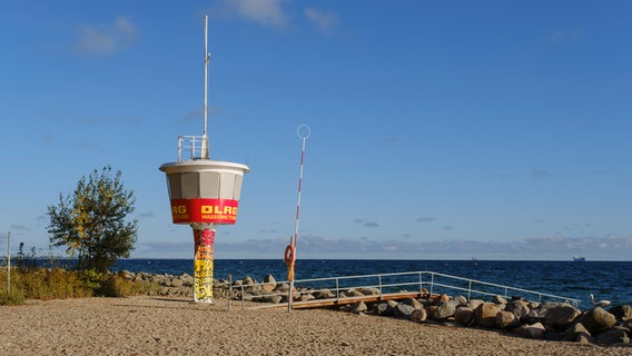 Ein DLRG-Aussichtsturm steht vor einem Steg einer Badestelle am Meer. Im Hintergrund sind am Horizont des Wassers Schiffe zu erkennen. © picture alliance / CHROMORANGE Foto: Wilfried Wirth