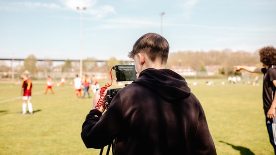 Ein junger Mann steht vor einer Videokamera und im Hintergrund läuft ein Fußballspiel. © NDR Foto: Lisa Pandelaki